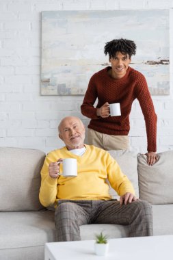 Smiling multiethnic grandfather and grandson holding cups and looking at camera at home 