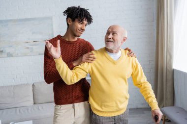 Cheerful grandparent with walking cane talking to african american grandson in living room 