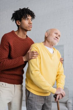 Young african american man hugging upset grandparent with walking cane at home