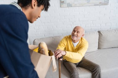 Blurred african american grandson holding package with food near smiling granddad at home 