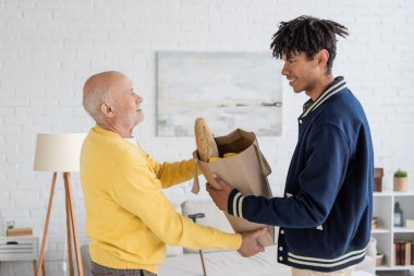 Side view of smiling interracial grandson and grandfather holding bag with food at home 