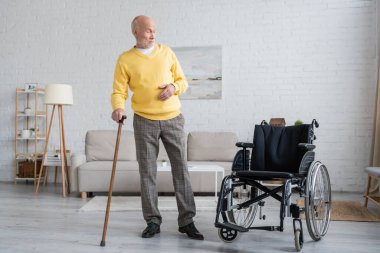 Elderly man holding walking cane and looking at wheelchair at home 