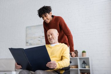 African american man standing near granddad with photo album in wheelchair at home 
