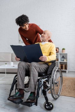 Positive pensioner holding photo album while sitting in wheelchair near african american grandson at home 