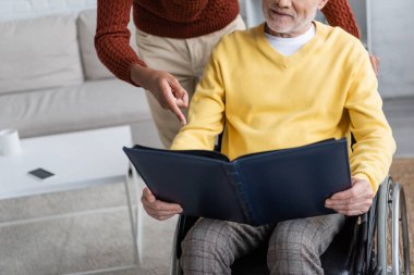 Cropped view of african american grandson pointing with finger near granddad with photo album in wheelchair at home 
