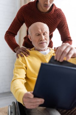 African american man hugging granddad in wheelchair holding blurred photo album at home 