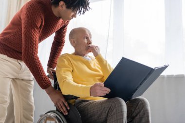 African american grandson looking at sad grandpa holding photo album in wheelchair at home 