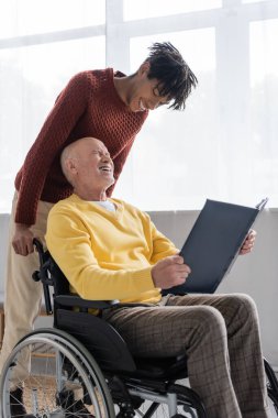 Positive pensioner holding photo album while sitting in wheelchair near african american grandson at home 
