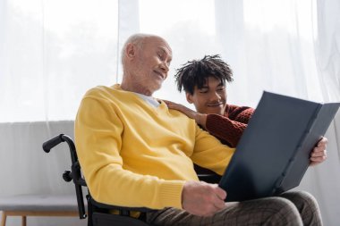 Smiling pensioner in wheelchair holding photo album near african american grandson at home 