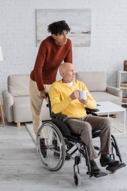 African american man standing near granddad holding cup in wheelchair at home 