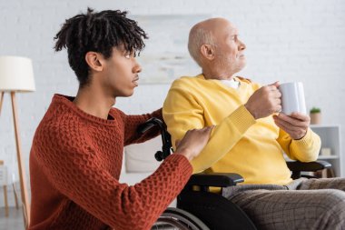Side view of african american grandson hugging grandfather with cup in wheelchair at home 