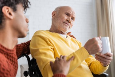 Sad senior man holding cup in wheelchair near blurred african american grandson at home 