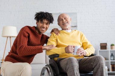 Smiling african american grandson hugging granddad with cup in wheelchair at home 