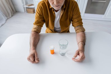 Cropped view of pensioner siting near pills and glass of water in kitchen 