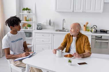 African american volunteer writing on clipboard near senior man with croissant and tea at home 