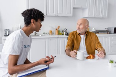 African american volunteer holding clipboard near senior man with cup in kitchen 