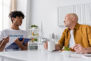 African american volunteer holding clipboard near senior man with cup at home 
