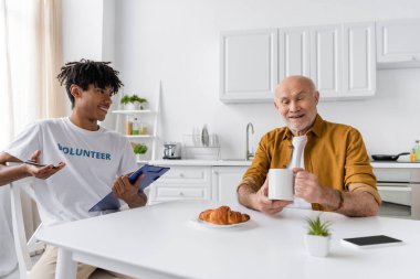 Smiling pensioner holding cup near african american volunteer at home 