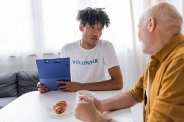 African american volunteer holding clipboard near blurred pensioner with cup at home 