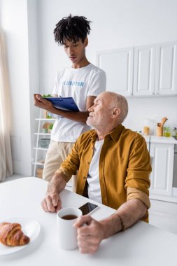 African american volunteer looking at clipboard near elderly man with tea at home 