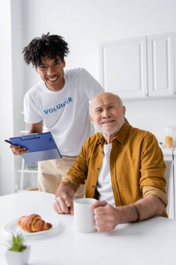 Smiling african american volunteer holding clipboard near elderly man with cup in kitchen 