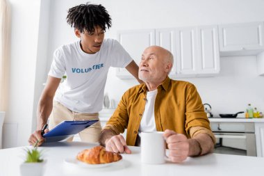African american volunteer holding clipboard near elderly man with cup and croissant at home 
