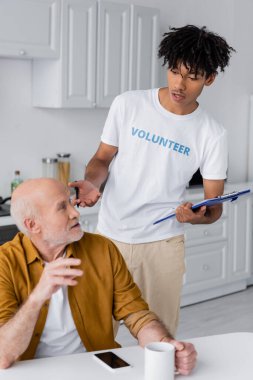 African american volunteer holding clipboard near pensioner with cup at home 