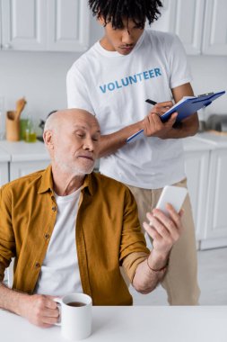 Senior man holding smartphone and cup near african american volunteer with clipboard at home 