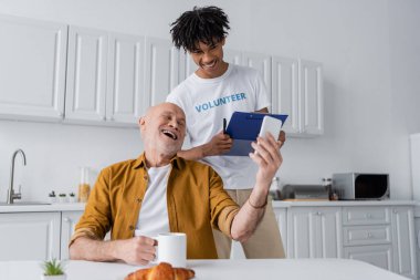 Positive senior man holding cellphone and cup near african american volunteer with clipboard in kitchen 