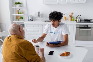 African american volunteer with clipboard talking to senior man with tea in kitchen 