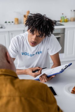 African american volunteer holding clipboard near blurred elderly man in kitchen 