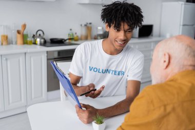 Cheerful african american volunteer pointing at clipboard near blurred senior man in kitchen 