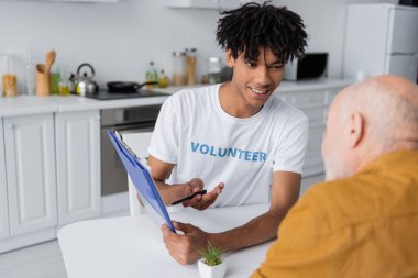 Cheerful african american volunteer pointing at clipboard near blurred senior man in kitchen  clipart