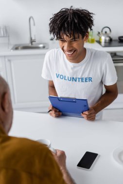Cheerful african american volunteer with clipboard looking at blurred senior man with tea near smartphone in kitchen 