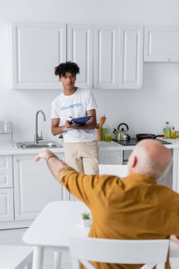 African american volunteer writing on clipboard near blurred pensioner in kitchen 