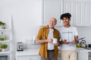 Positive senior man holding cup near african american volunteer with clipboard in kitchen 