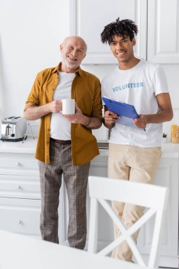 Smiling african american volunteer with clipboard and pensioner with cup looking at camera in kitchen 