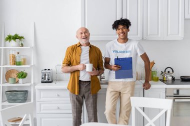 Cheerful african american volunteer with clipboard and elderly man looking at camera in kitchen 