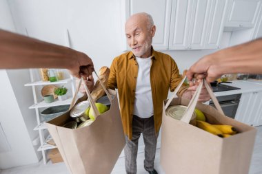 Smiling pensioner taking bags with food from african american volunteer at home 