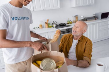 Smiling african american volunteer holding package with food near pensioner in kitchen 