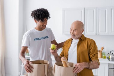 Smiling african american volunteer standing near pensioner and packages with food in kitchen 