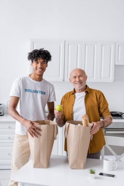 Positive african american volunteer standing near elderly man and bags with food in kitchen 