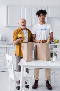 Smiling senior man and african american volunteer looking at camera near food in bags in kitchen 