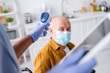 African american nurse holding pyrometer and digital tablet near blurred senior an in medical mask at home 