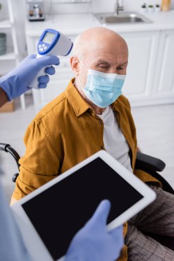 African american nurse holding pyrometer and digital tablet near pensioner in medical mask and wheelchair at home 