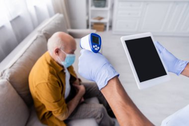 African american nurse holding pyrometer and digital tablet with blank screen near blurred pensioner in medical mask at home 