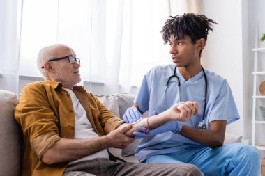 African american nurse in latex gloves holding cotton pad on hand of senior patient at home 