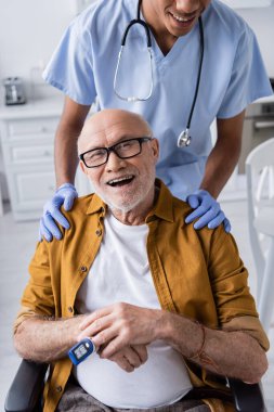 Cheerful elderly man with pulse oximeter looking at camera near african american nurse at home 