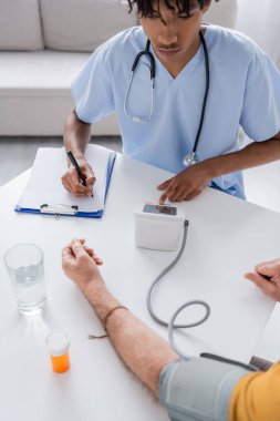 African american nurse writing on clipboard near tonometer and senior patient at home 
