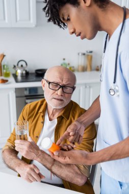 African american nurse pointing at pills near senior patient with glass of water at home 